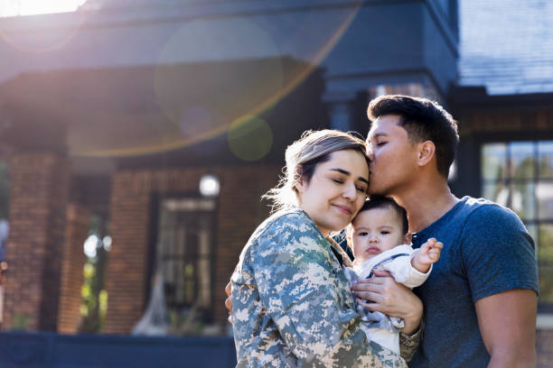 While standing in their front yard, a mid adult husband kisses his soldier wife before she leaves for an assignment. The woman is holding their baby girl.